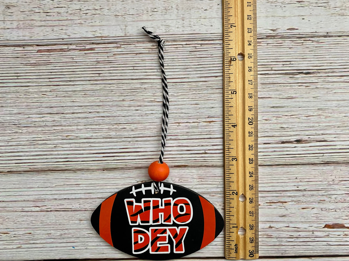 A football-shaped hanging ornament with the phrase 'WHO Dey' in red and white lettering, black and orange stripes, and a black-and-white string, shown next to a wooden ruler on a whitewashed wood background.