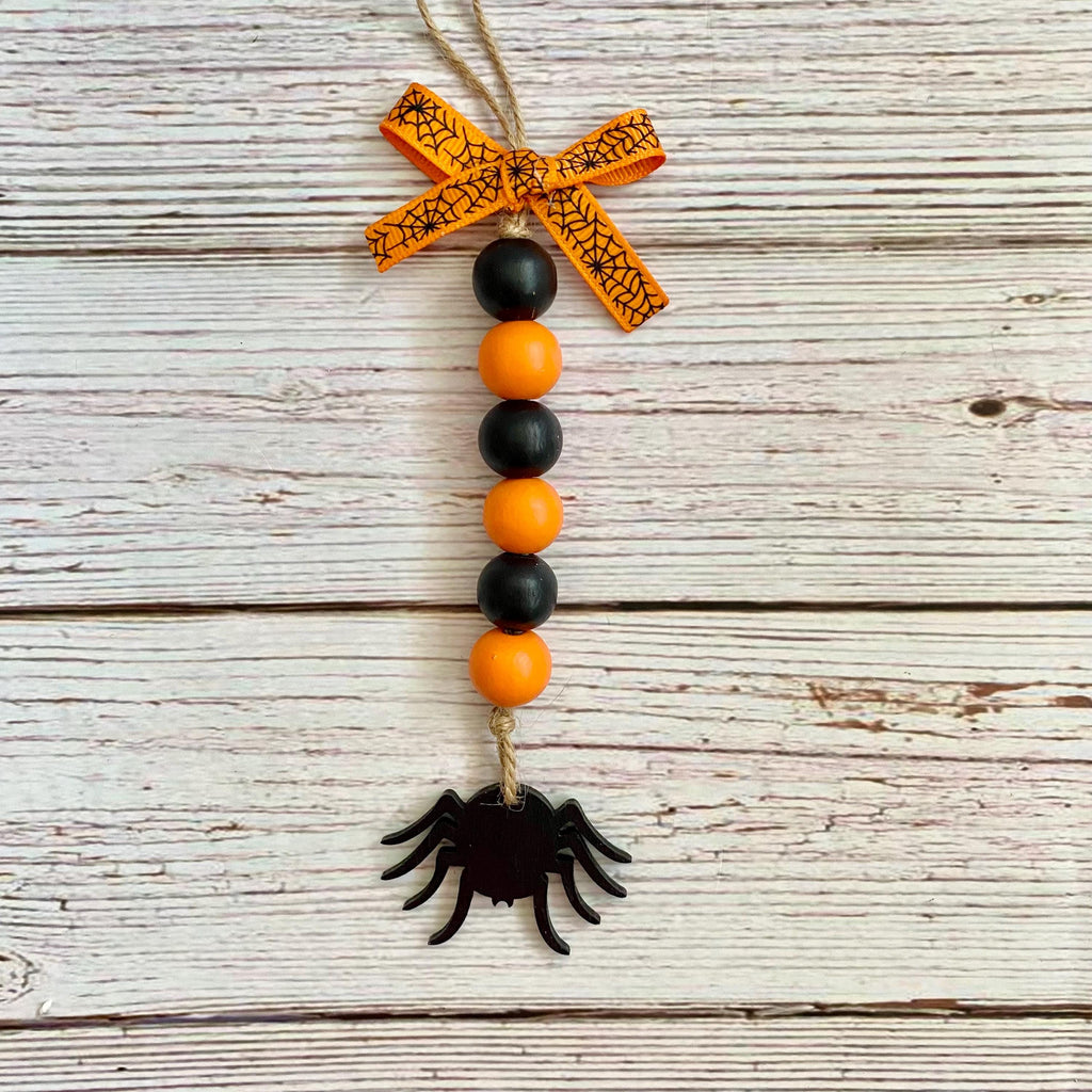 Halloween beaded hanging ornament with orange and black beads, an orange spider silhouette, and an orange patterned ribbon bow at the top, shown on a whitewashed wood plank background.