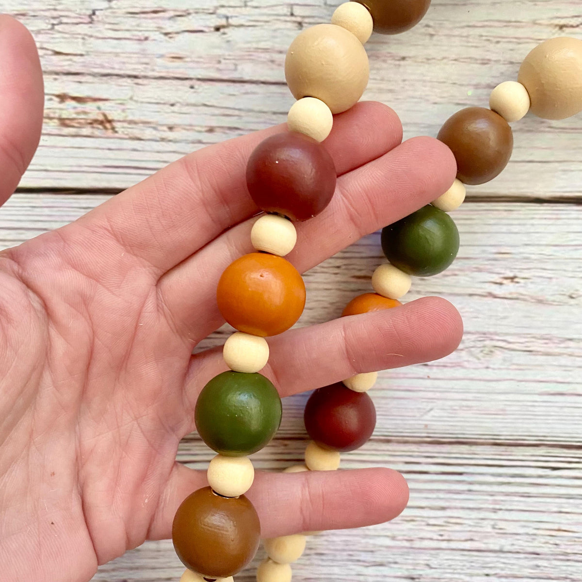 A close-up of a hand holding a wooden bead garland with alternating light cream, medium brown, dark reddish-brown, and olive green round beads, set against a rustic white-washed wood plank background.