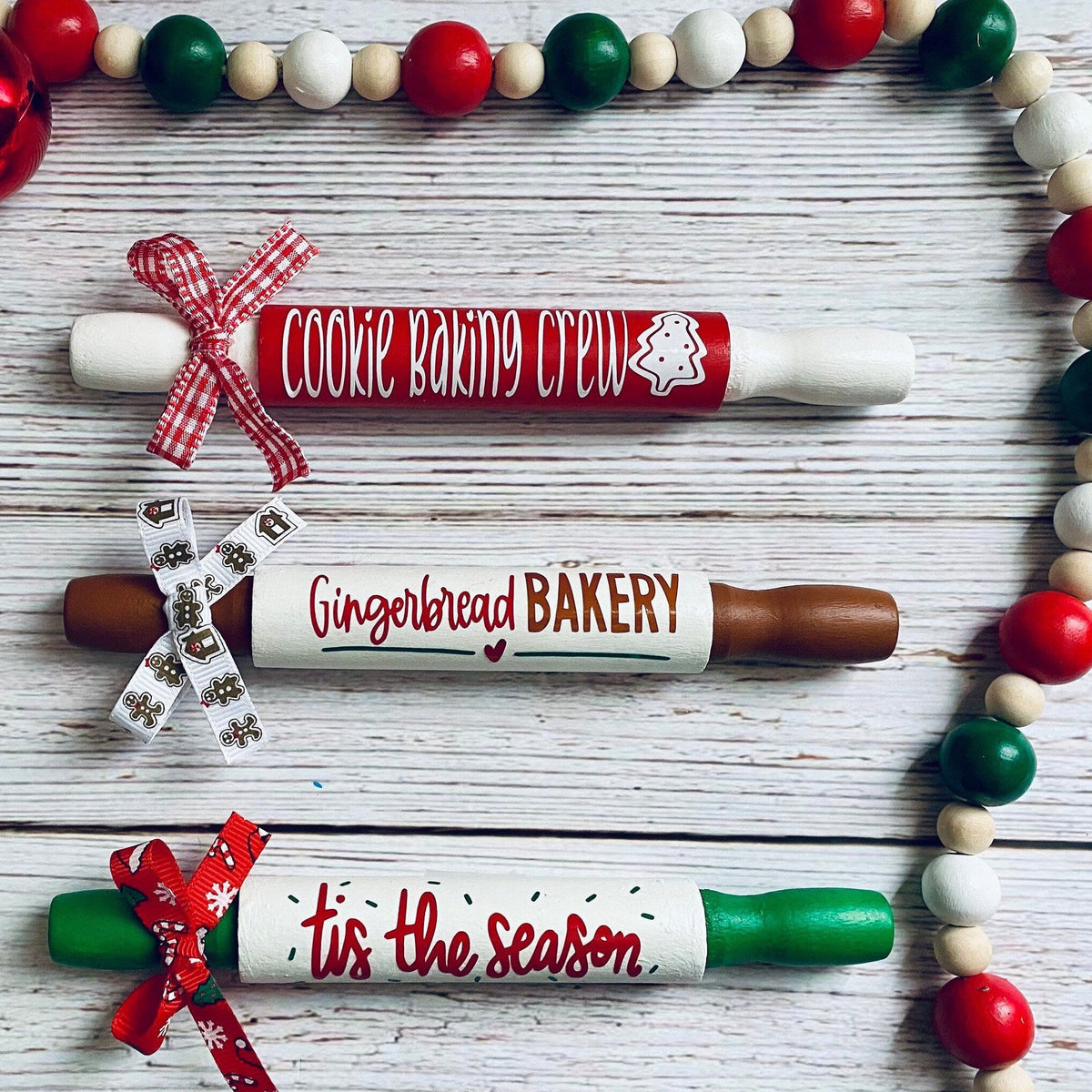 Three Christmas-themed wooden rolling pins with personalized baking and holiday messages, styled on a white wood background with a red and green beaded garland.