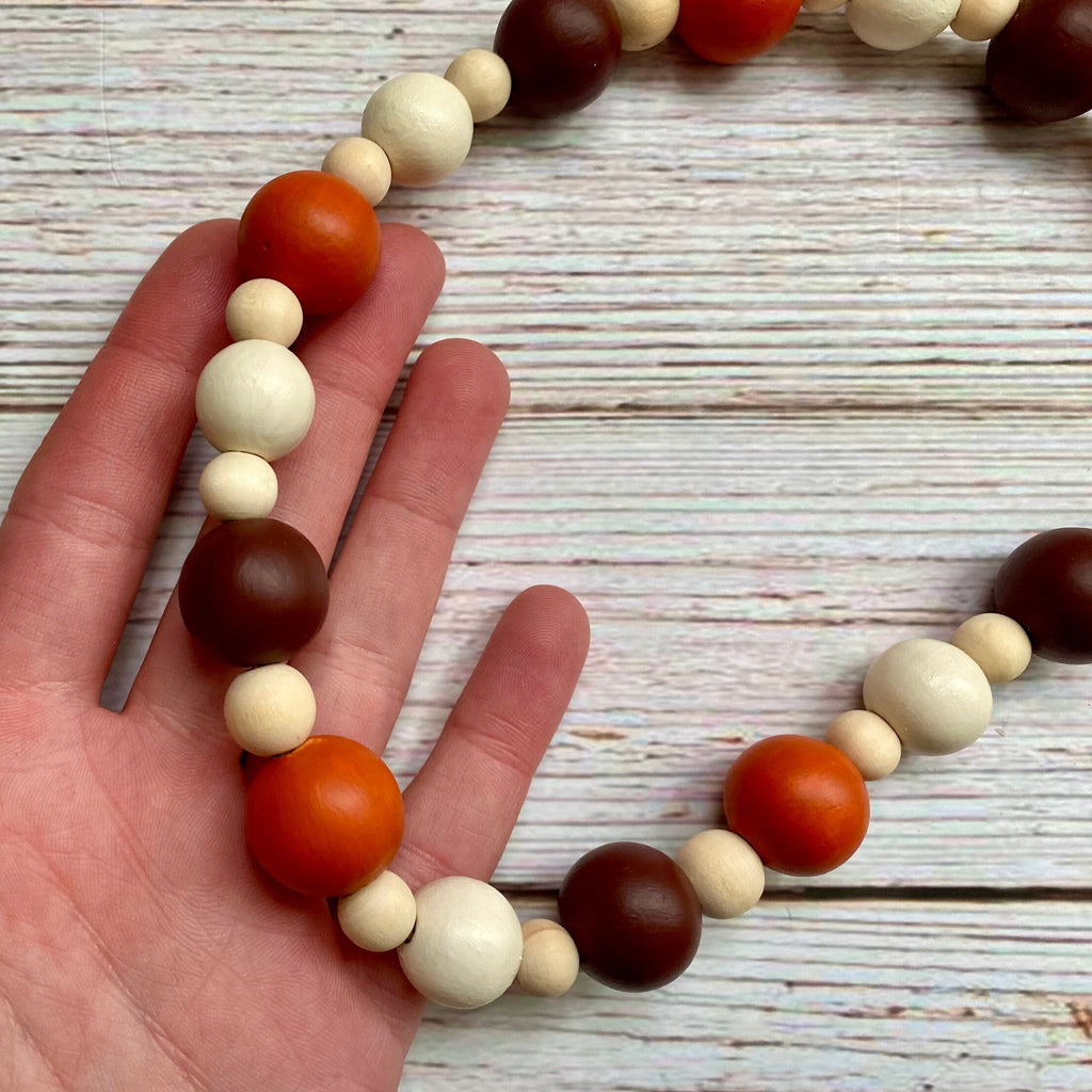 A close-up of a hand holding a beaded necklace with alternating matte orange, cream, and dark brown wooden beads, set against a rustic white-washed wood background.