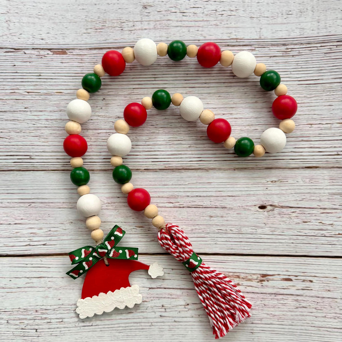 A Christmas-themed wooden bead garland with red, green, and white beads, a red Santa hat charm with a green patterned ribbon, and a red-and-white striped tassel, displayed on a light gray wood plank background.