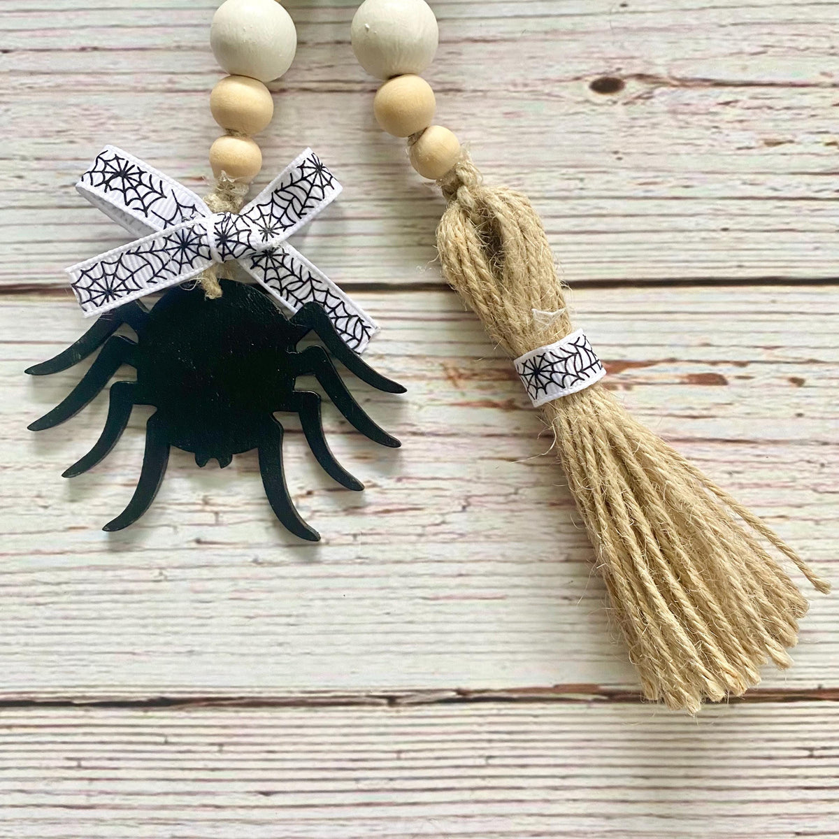 Two Halloween-themed beaded garland ornaments on a whitewashed wooden background. One ornament features a black spider silhouette hanging from a white ribbon with a black spiderweb pattern.