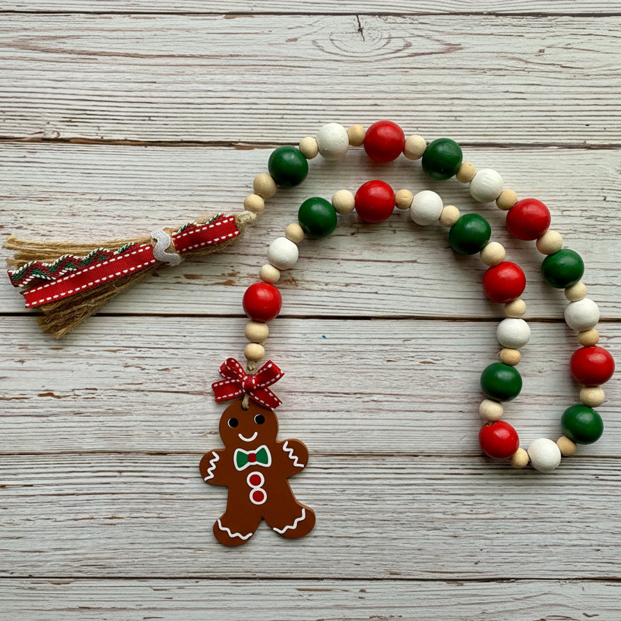 A Christmas-themed beaded garland with red, green, and white wooden beads, a red and green ribbon tassel, and a gingerbread man ornament, displayed on a rustic white-washed wooden background.