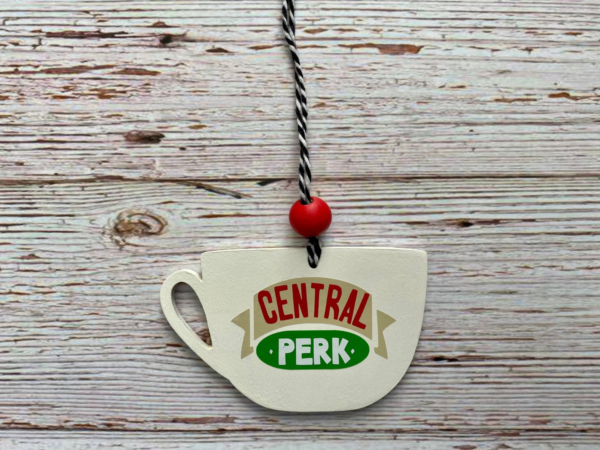 A ceramic teacup-shaped ornament hanging from a black-and-white string with a red bead, featuring the 'Central Perk' logo from Friends on a distressed white wood background.