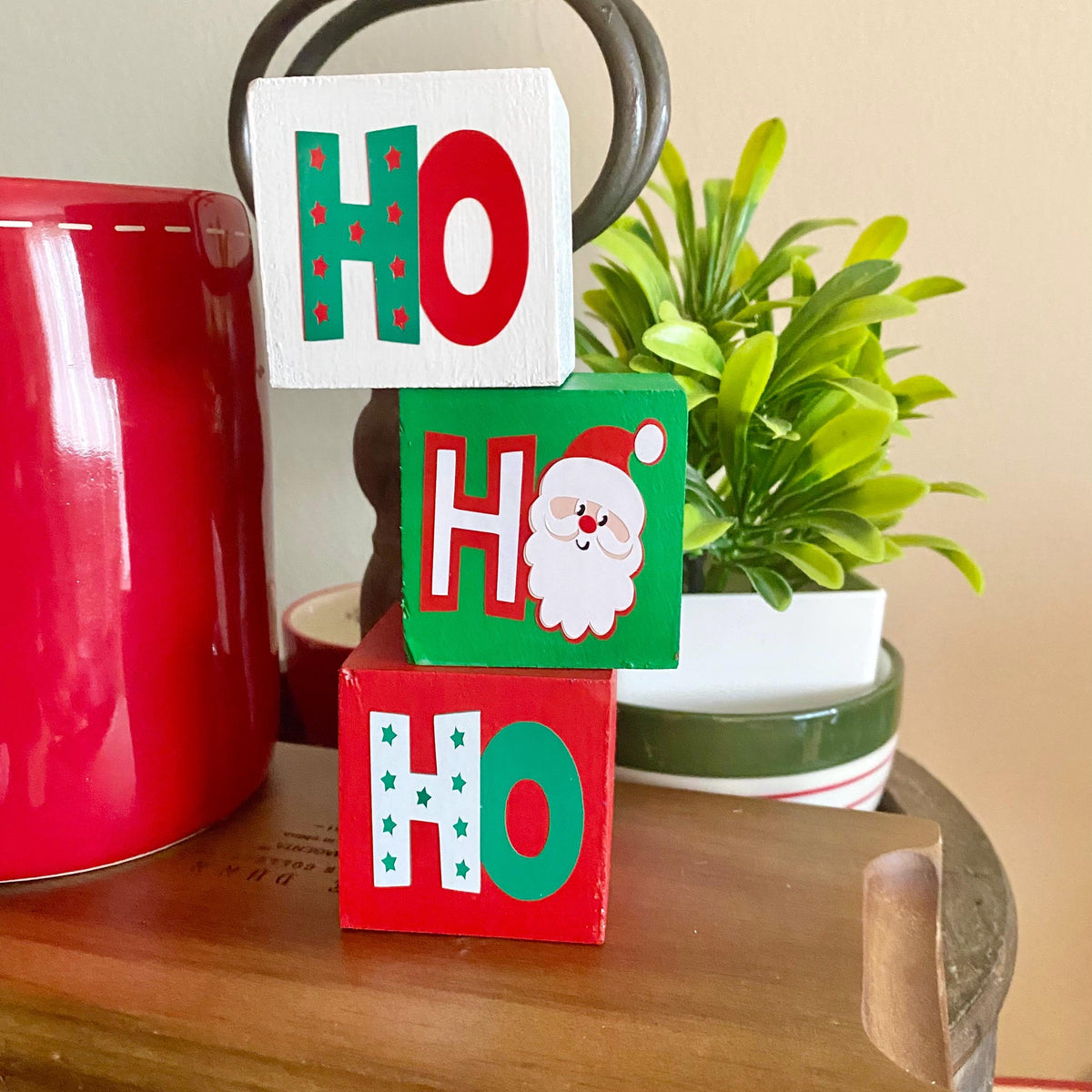 Three stacked wooden HO blocks with Christmas-themed lettering and a Santa face, displayed on a wooden table next to a red mug and a potted plant.
