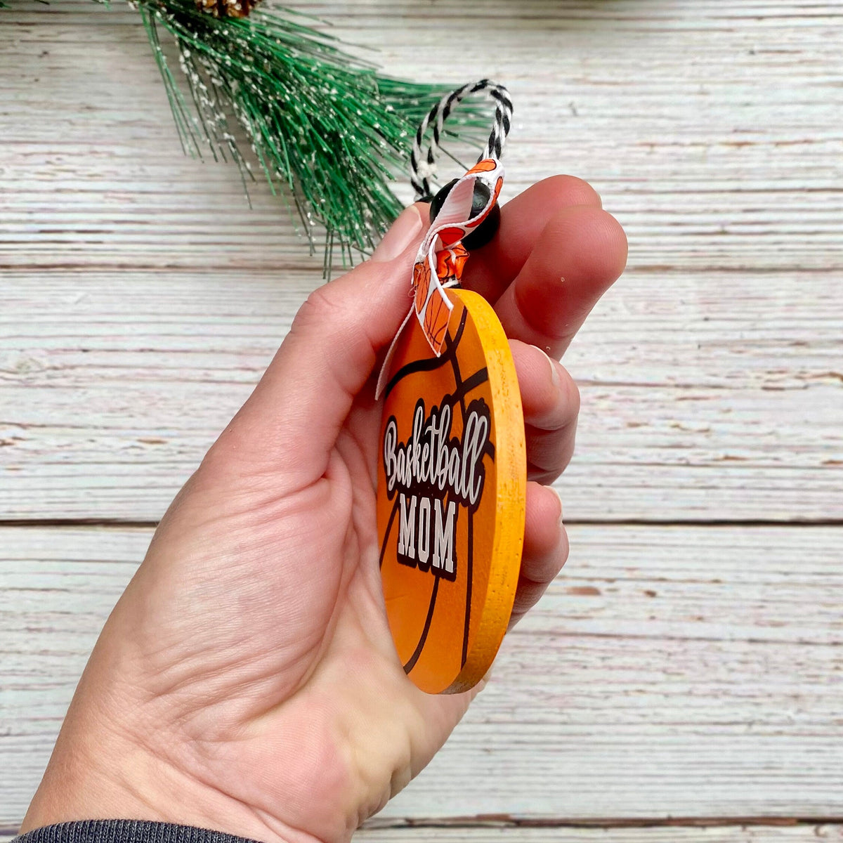 A hand holding a round orange basketball-themed ornament with the words 'Basketball Mom' printed on it, against a rustic white wood plank background.