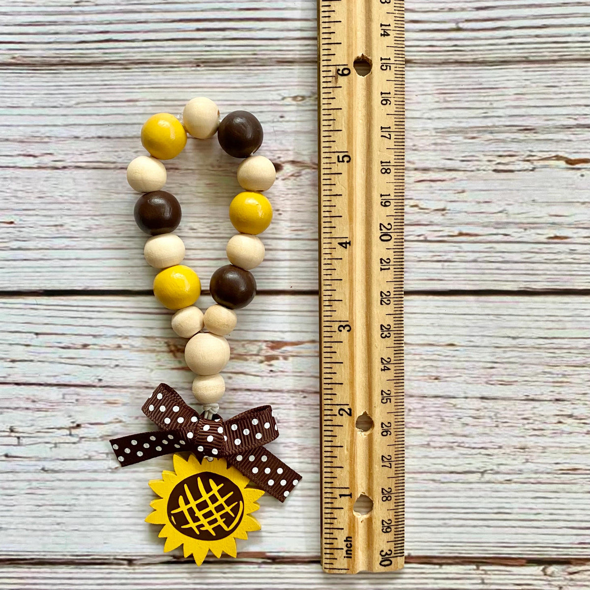 A wooden bead garland with yellow, brown, and cream beads, a brown polka dot ribbon bow, and a yellow sunflower charm, displayed next to a wooden ruler on a white-washed wood background.