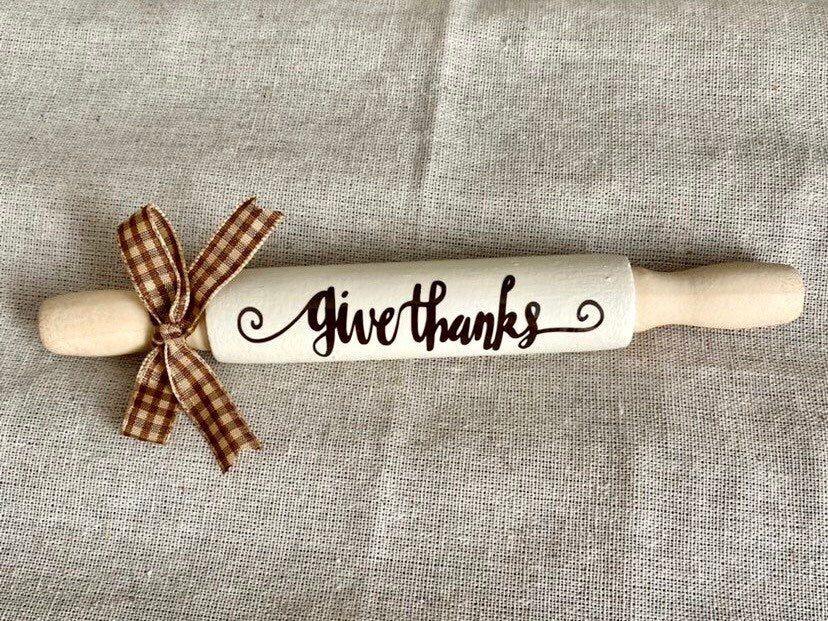 A wooden rolling pin with 'Give thanks' written on it in black script, tied with a brown and beige plaid ribbon, set against a textured linen background.