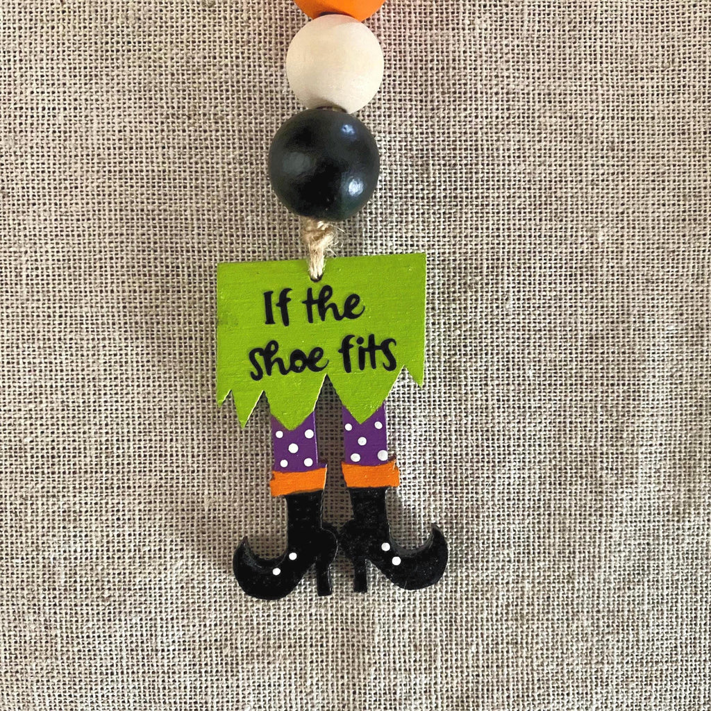 A Halloween-themed wooden bead garland charm shaped like a witch's legs, with the text 'If the shoe fits' on a green sign. The charm features purple polka-dot pants, orange cuffs, black shoes, and is suspended from wooden beads on a beige linen background.