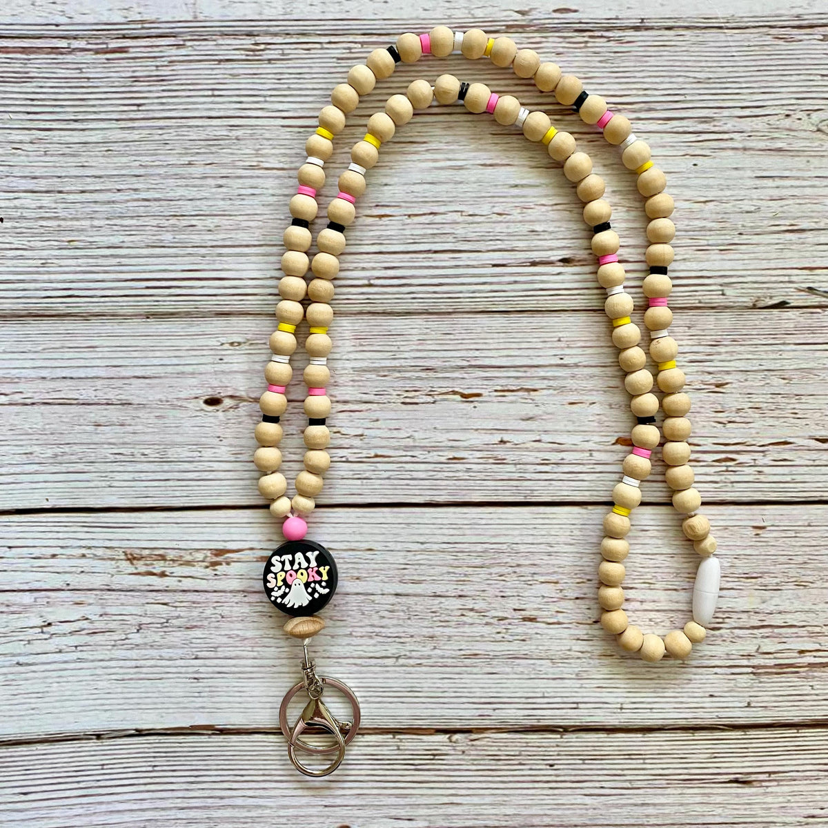 A beaded necklace with natural wood beads, small pink, yellow, and black accents, a round black charm reading 'STAY SPOOKY', and a silver keyring clasp, laid flat on a whitewashed wooden surface.
