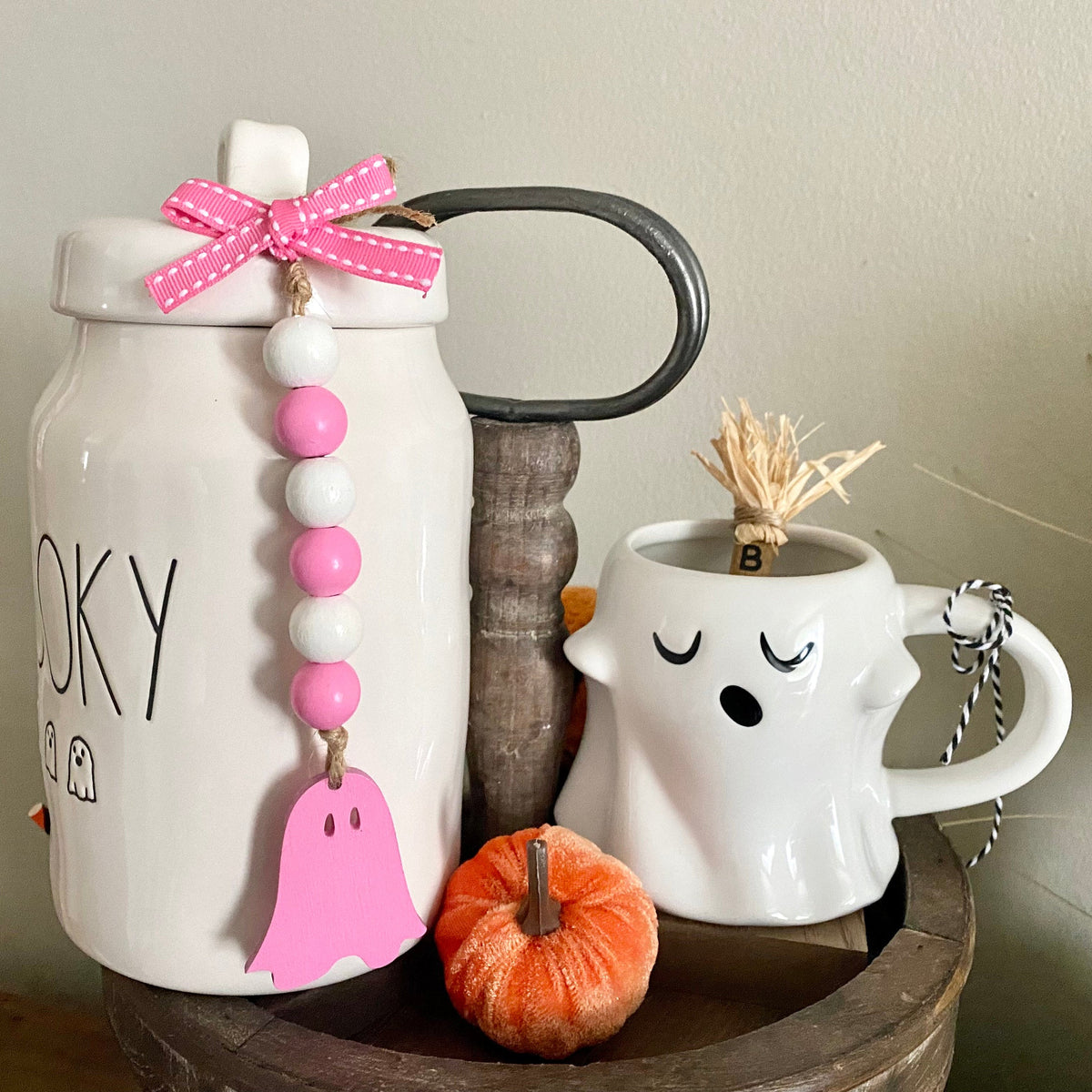A styled tabletop display featuring a white ceramic canister with a pink ghost charm and bow, next to a white ghost-shaped mug with a straw hat, a small orange pumpkin, and a wooden spool on a neutral background.