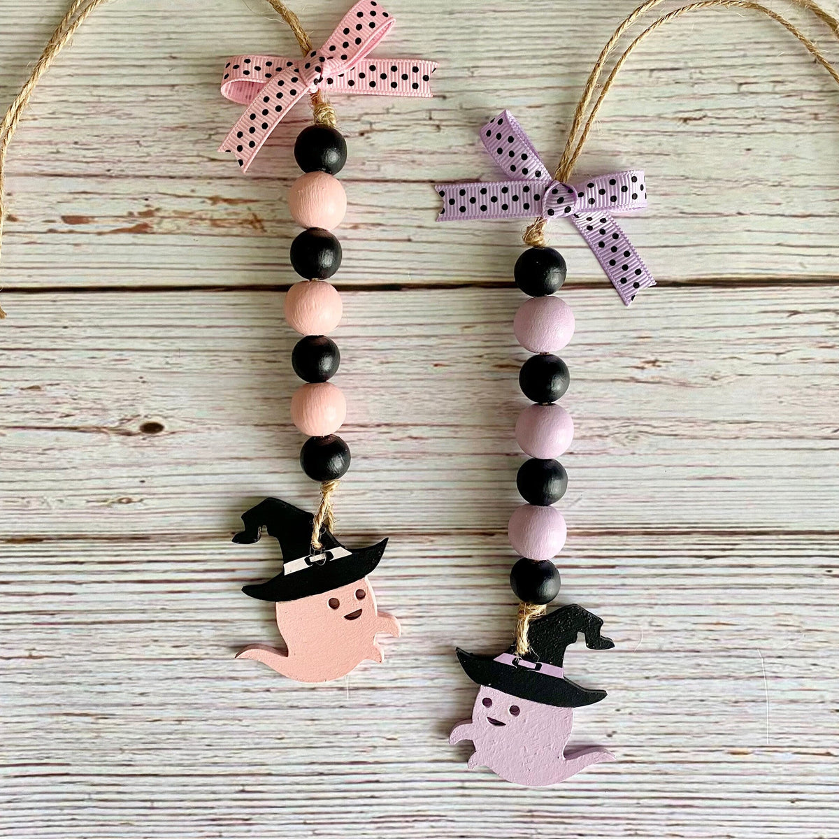 Two Halloween hanging ornaments with black and pastel pink wooden beads, pink and lavender polka-dot bows, and ghost and witch figures, displayed on a whitewashed wood background.
