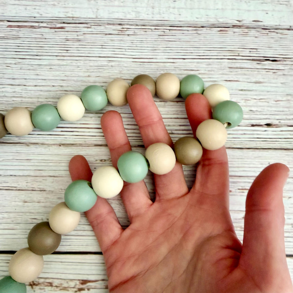 A hand holding a strand of matte wooden beads in pastel shades of sage green, cream, and taupe, displayed against a rustic white-washed wood background.