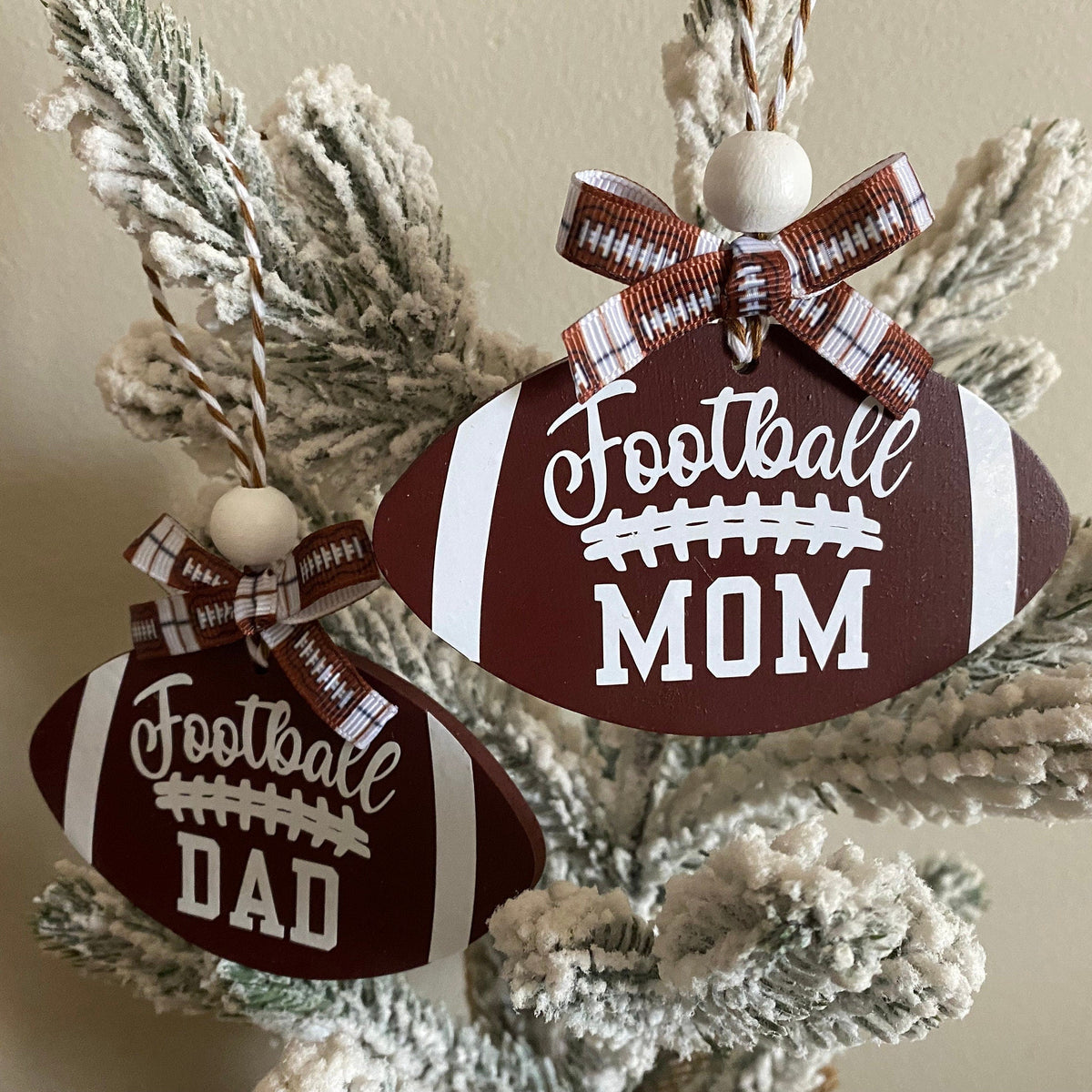 Two football-shaped Christmas ornaments hanging on a frosted tree. One ornament says "Football DAD" and the other says "Football MOM" in white lettering, both with brown and white striped bows.