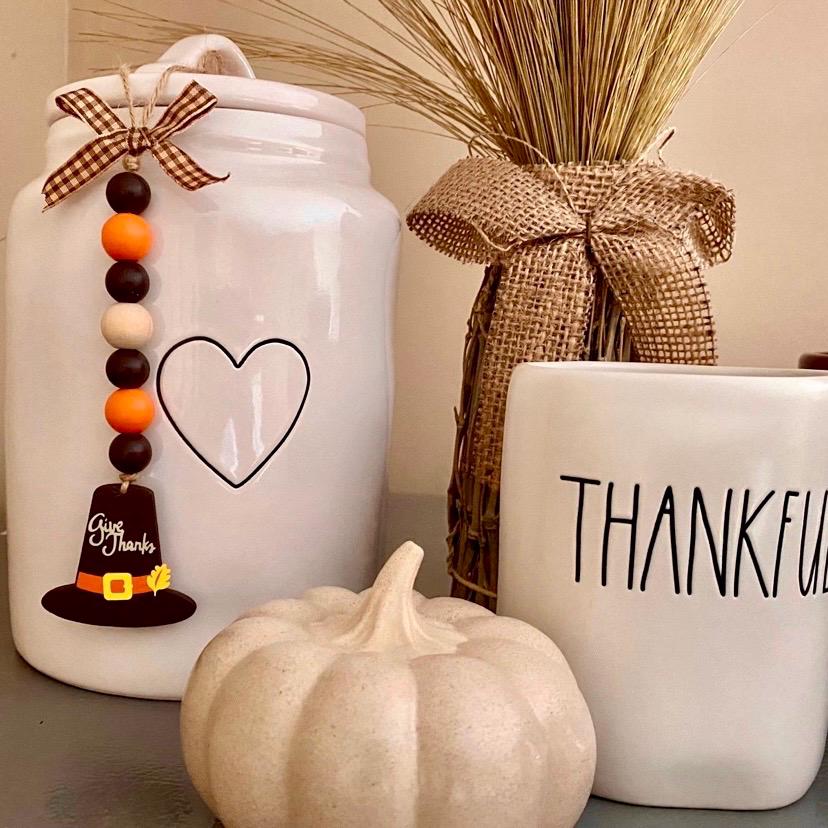 White ceramic Thanksgiving-themed decor set including a mason jar with a heart outline and hanging pilgrim hat charm, a white mug with partial 'THANKFUL' text, a small cream pumpkin, and a burlap-wrapped decorative bundle in the background.