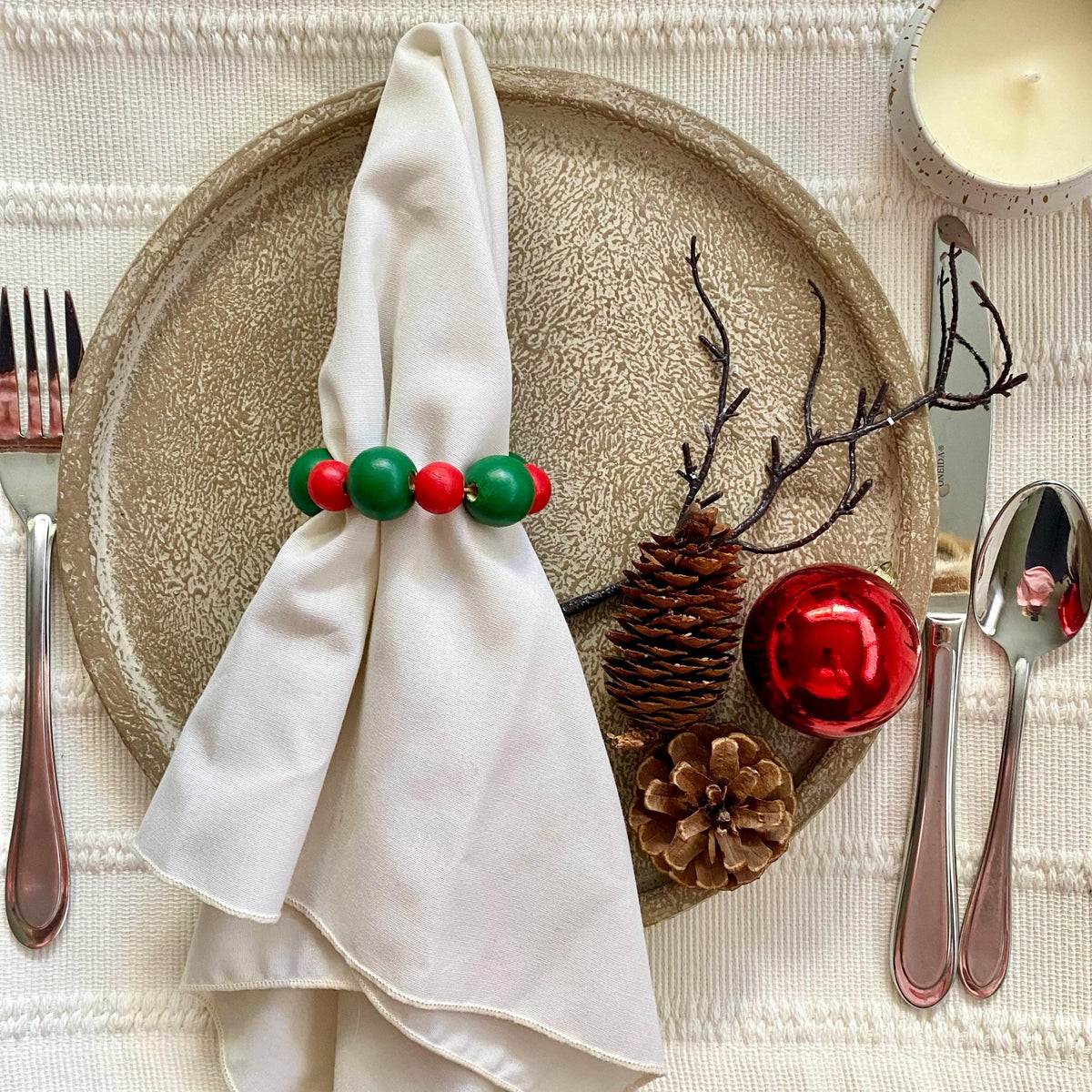 A styled holiday table setting featuring a cream-colored textured placemat, a round beige charger plate, a cream napkin held by a red and green beaded ring, pinecones, a red ornament, and silver flatware.