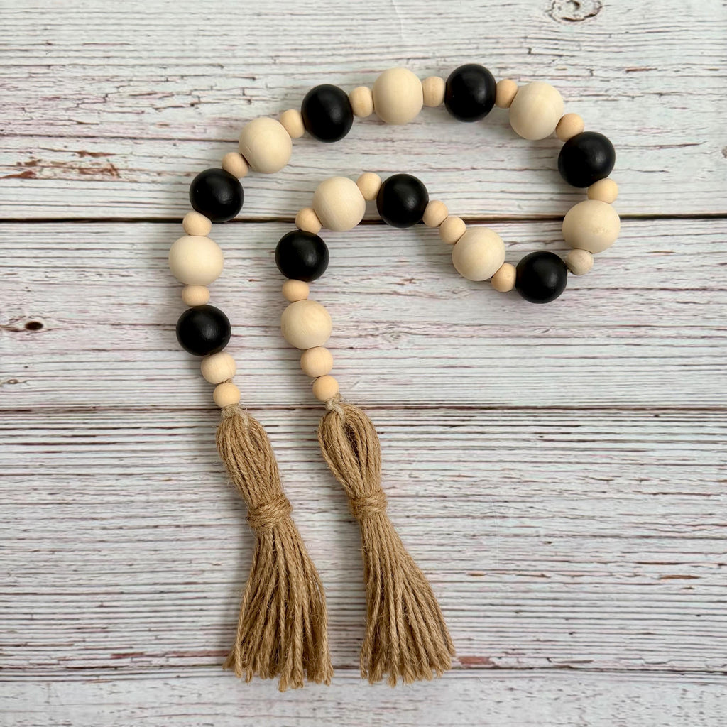A bohemian-style wooden bead garland with alternating matte black and natural light wood beads, finished with two knotted jute tassels, displayed on a white-washed wooden background.
