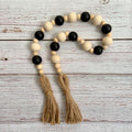 A bohemian-style wooden bead garland with alternating matte black and natural light wood beads, finished with two knotted jute tassels, displayed on a white-washed wooden background.