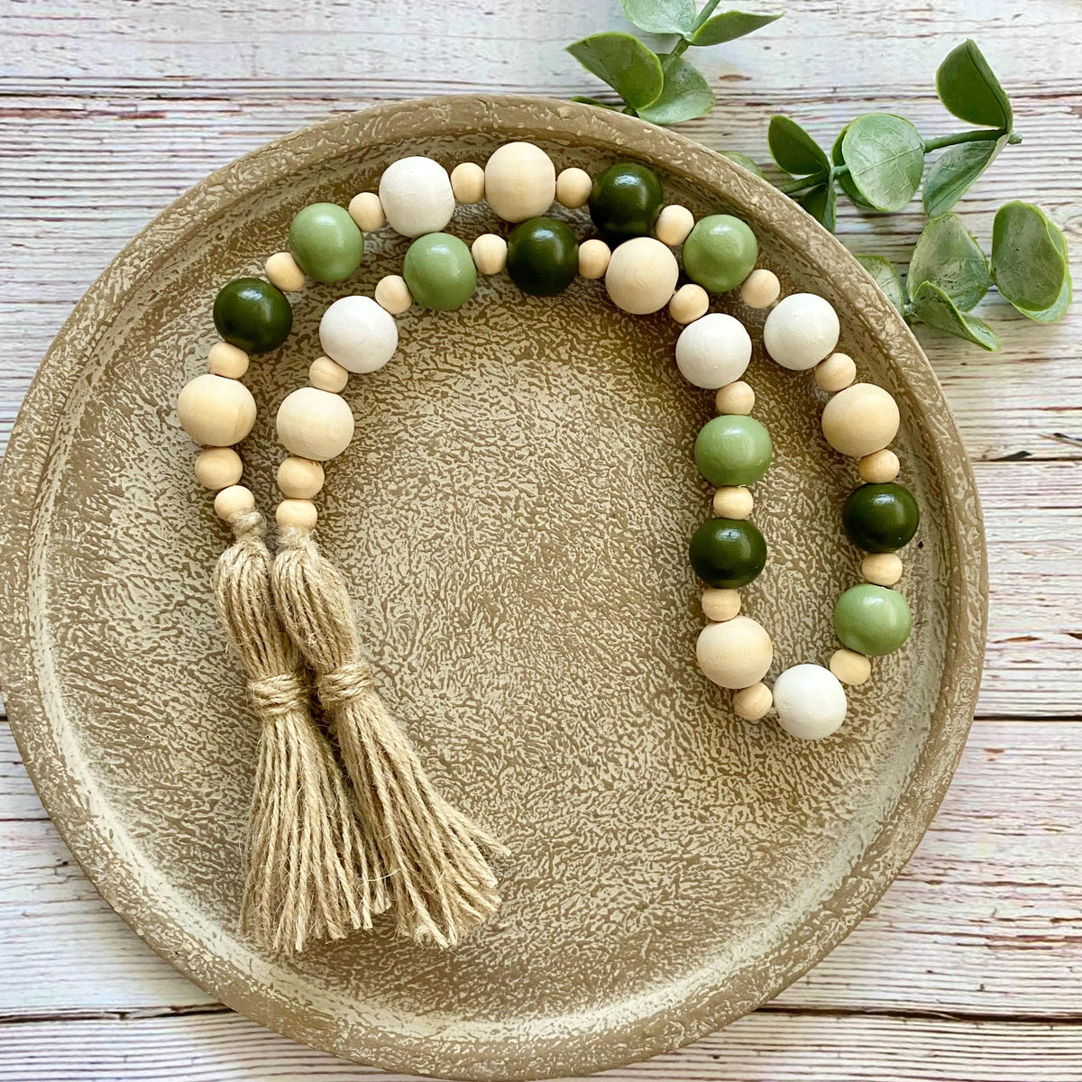 A beaded garland with natural wood beads, green wooden beads, and two beige tassels, displayed on a round textured gold tray with green leaves on a white wooden surface.