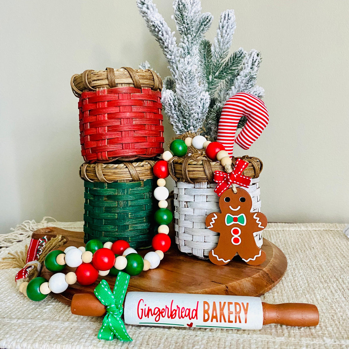 A styled holiday gingerbread bakery display featuring stacked woven baskets in red, green, and white with faux flocked pine branches, a red-and-white striped candy cane, a gingerbread cookie decoration, colorful bead garlands, and a white rolling pin labeled 'Gingerbread BAKERY' on a wooden board.