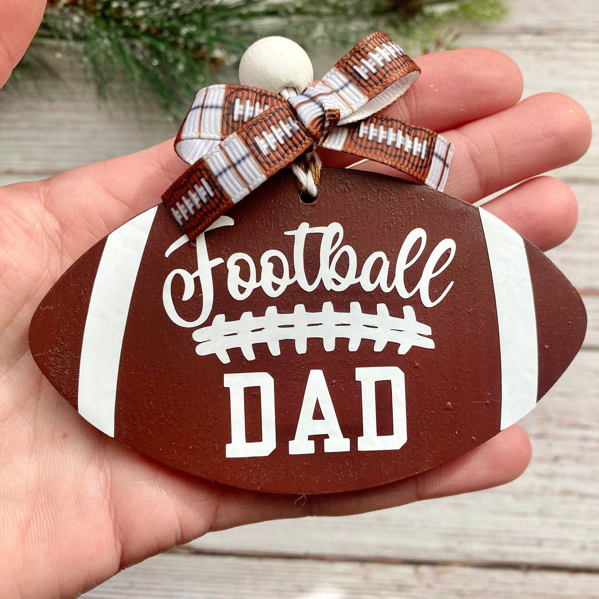 A hand holding a football-shaped wooden ornament with 'Football DAD' written in white, decorated with a brown and white plaid ribbon bow.