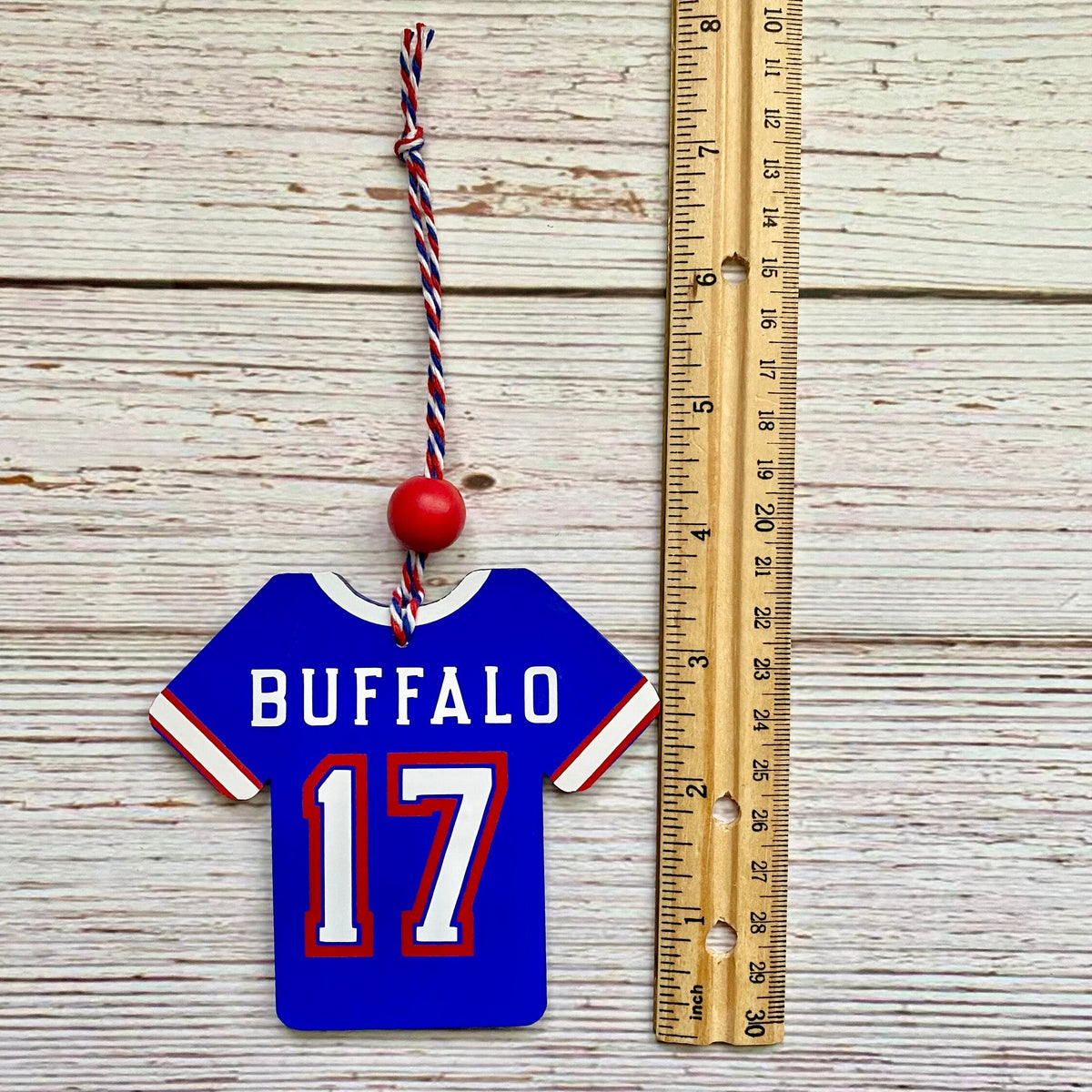 A small wooden Buffalo jersey ornament with the number 17 and a red hanging cord, shown next to a wooden ruler on a whitewashed wood plank background.