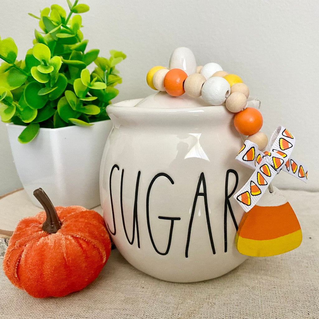 A ceramic sugar bowl with the word 'SUGAR' in black script, decorated with a wooden bead garland and a small paper windmill charm. It is styled on a beige fabric surface next to a small orange pumpkin and a green potted plant.