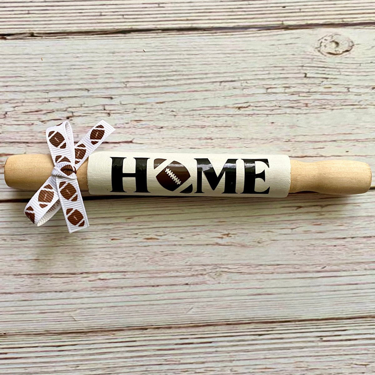 A wooden rolling pin styled as a decorative wall sign with 'HOME' written on the handle, featuring a football graphic and a white ribbon bow with football patterns, set on a rustic light wood plank background.