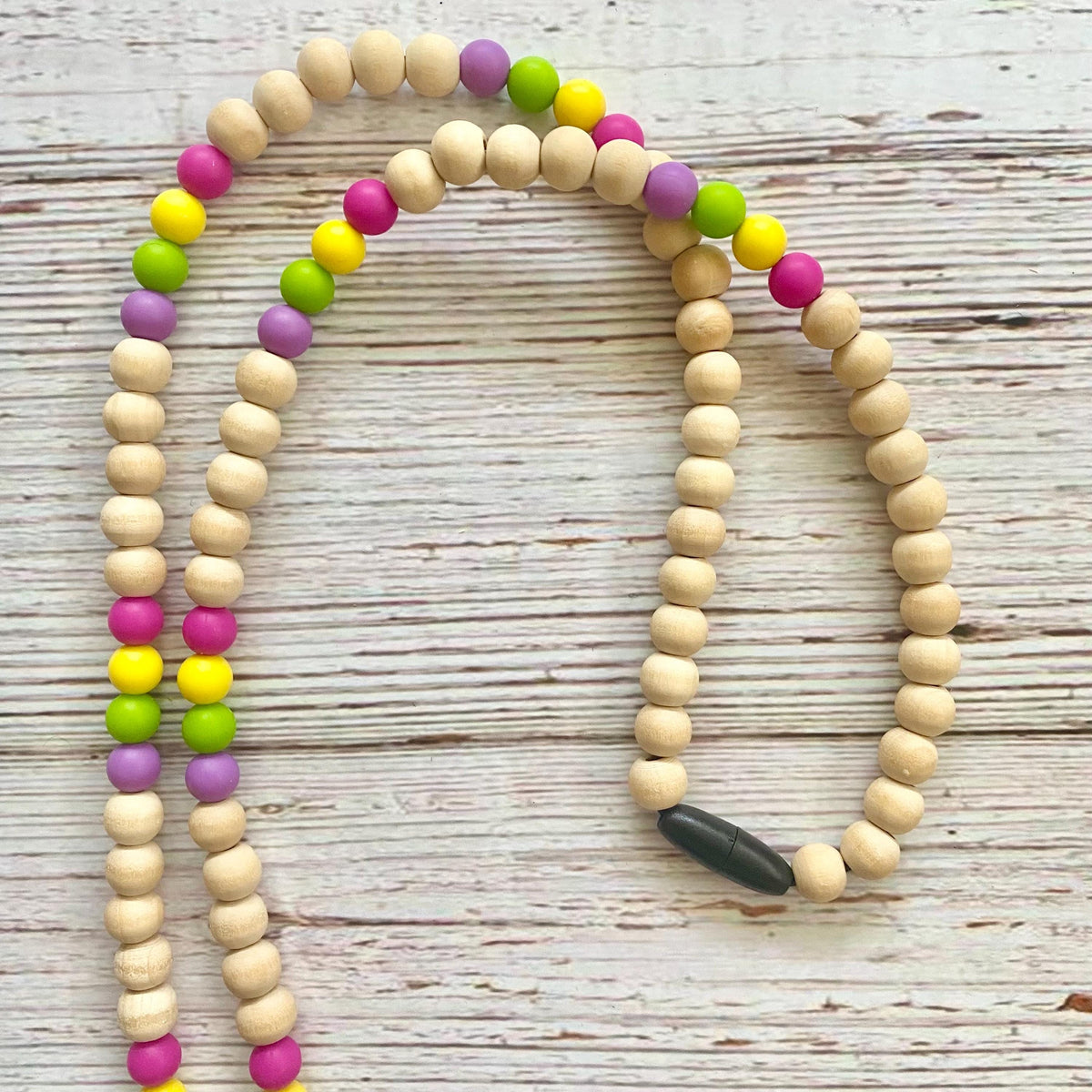 A beaded necklace made of natural light wood beads with alternating colorful accent beads in pink, purple, green, and yellow, displayed on a whitewashed wooden surface.