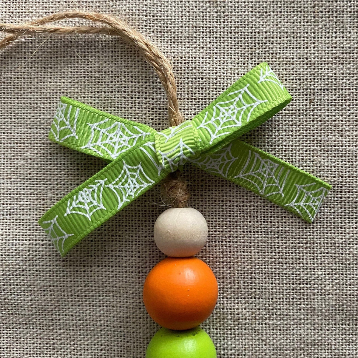 A handmade wooden bead garland ornament featuring a green and white spiderweb patterned ribbon bow, three stacked wooden beads in cream, orange, and light green, and a jute hanging string, displayed on a textured beige burlap background.
