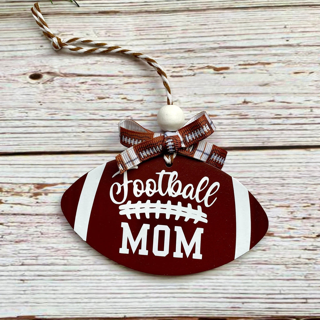 A football-shaped wooden hanging ornament with a brown and white striped ribbon bow, a white bead, and a twine loop. The ornament is dark red with white stripes and the words 'Football MOM' in white script and block lettering, shown on a whitewashed wood plank background.