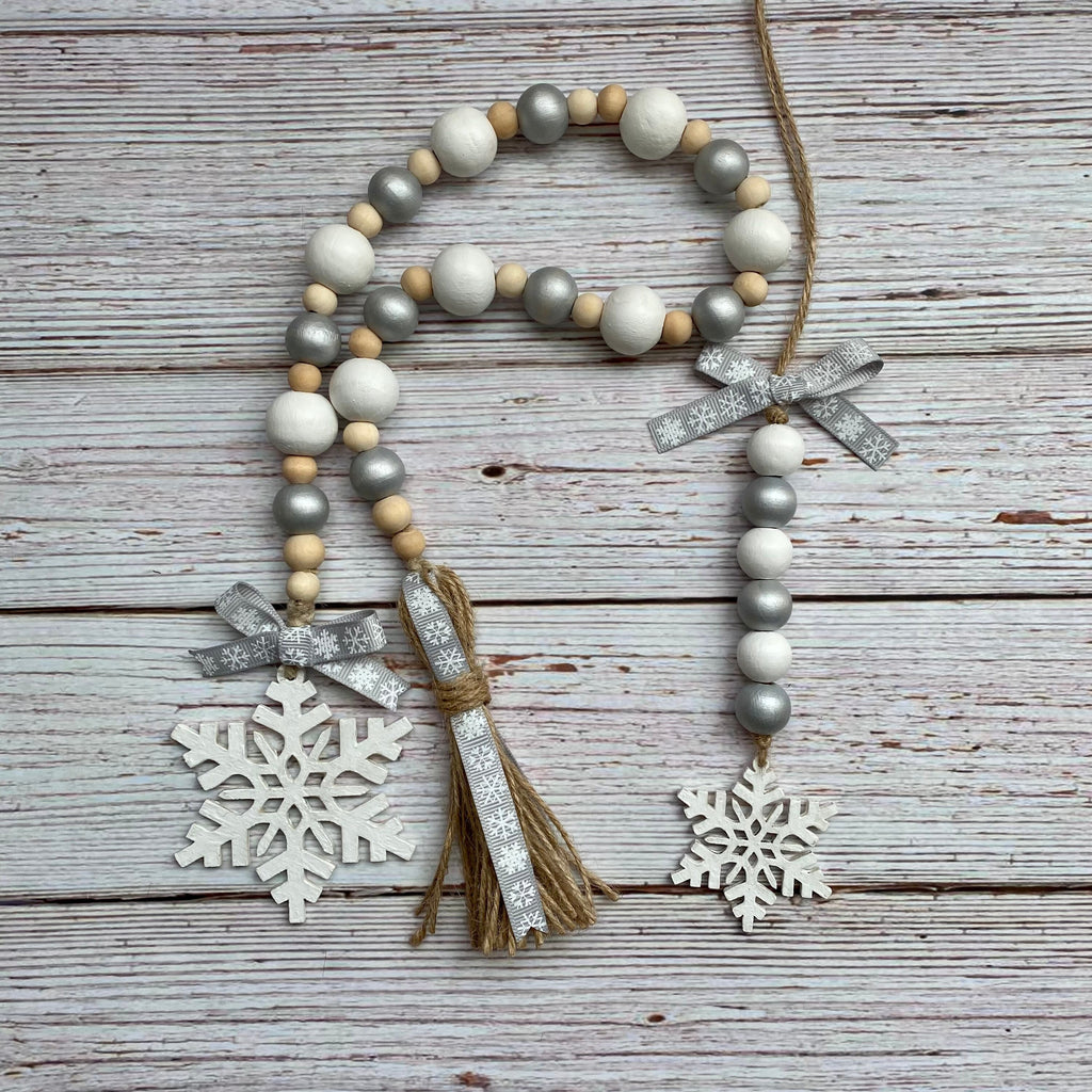 A rustic holiday garland made of white, gray, and natural wood beads, decorated with two silver glitter bows, a jute tassel, and two white wooden snowflakes, displayed on a weathered gray wood plank background.