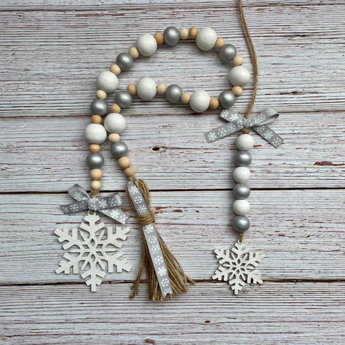 A rustic holiday garland made of white, gray, and natural wood beads, decorated with two silver glitter bows, a jute tassel, and two white wooden snowflakes, displayed on a weathered gray wood plank background.