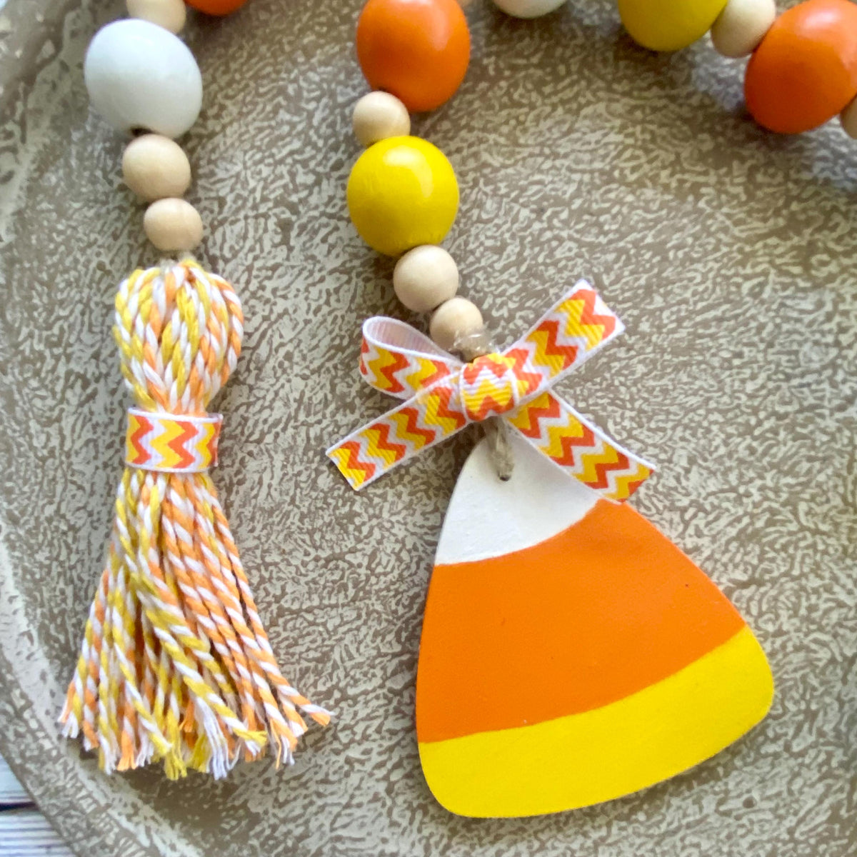 A close-up of a decorative beaded strand with wooden beads, a white tassel wrapped in orange and yellow chevron ribbon, and a candy corn-shaped charm tied with a matching ribbon, set on a textured beige surface.