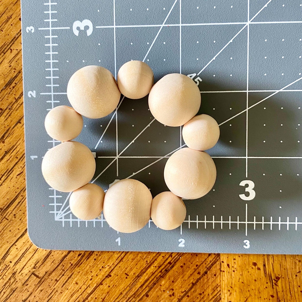 A set of nine natural wood beads in various sizes arranged in a circle on a gray cutting mat with grid lines, placed on a wooden table.