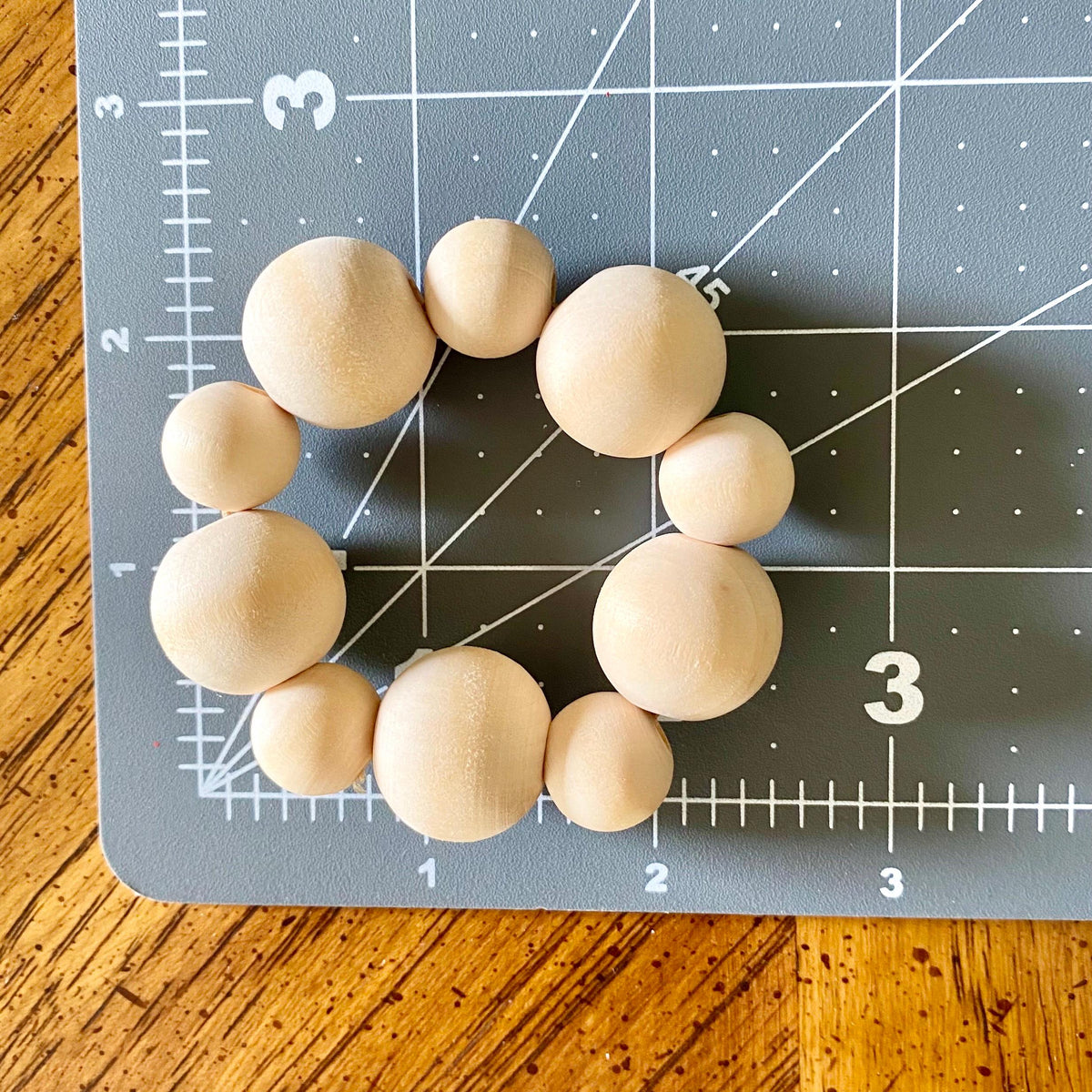 A set of nine natural wood beads in various sizes arranged in a circle on a gray cutting mat with grid lines, placed on a wooden table.