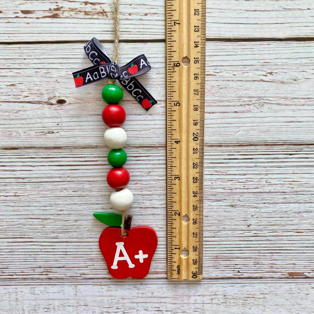 A decorative hanging ornament shaped like a red apple with green leaves, a black ribbon printed with the word 'ACB', and colorful wooden beads, shown next to a wooden ruler on a whitewashed wood plank background.