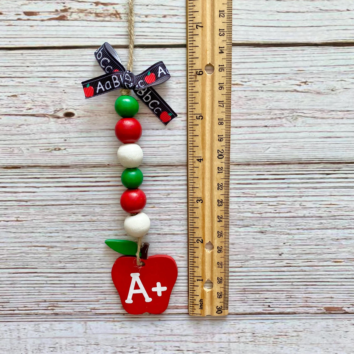 A decorative hanging ornament shaped like a red apple with green leaves, a black ribbon printed with the word 'ACB', and colorful wooden beads, shown next to a wooden ruler on a whitewashed wood plank background.