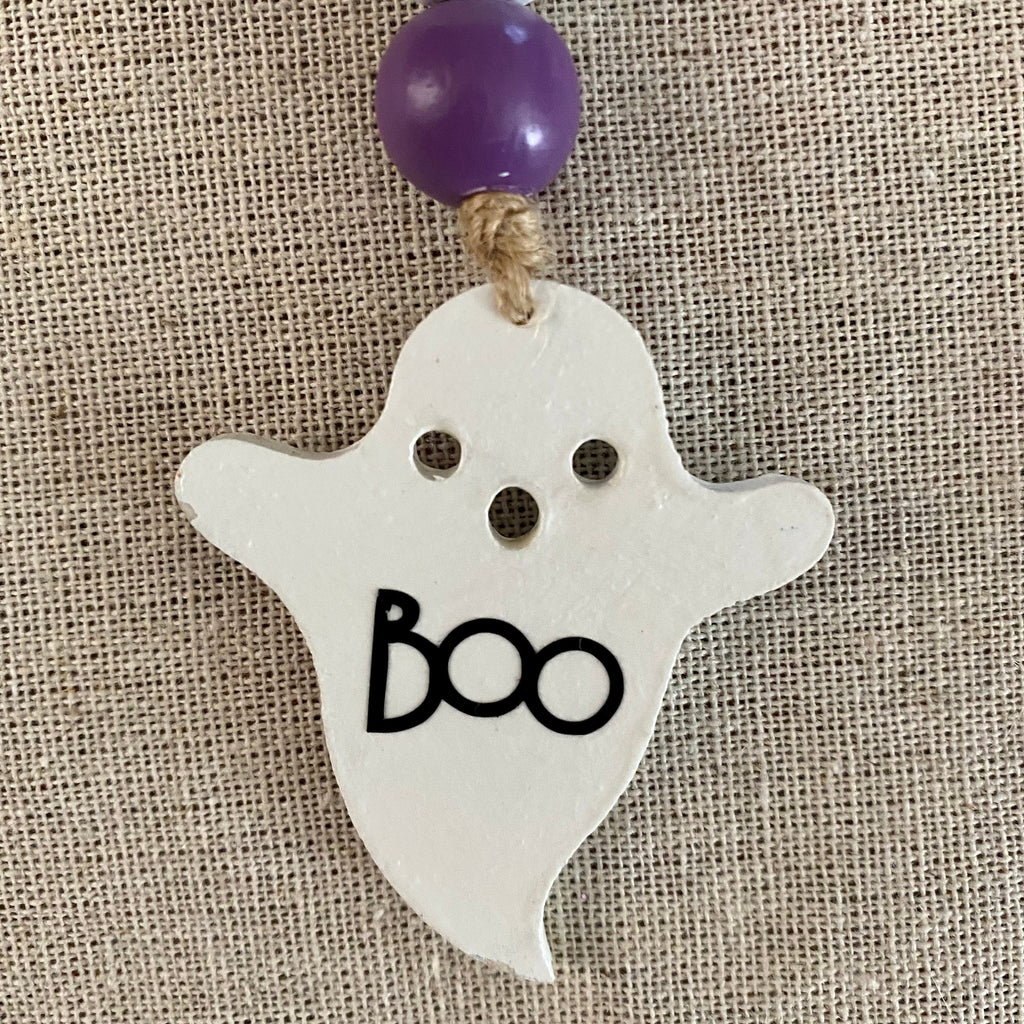 A white ghost-shaped hanging ornament with black 'BOO' lettering, two eye cutouts, and a purple bead at the top, shown against a beige burlap background.