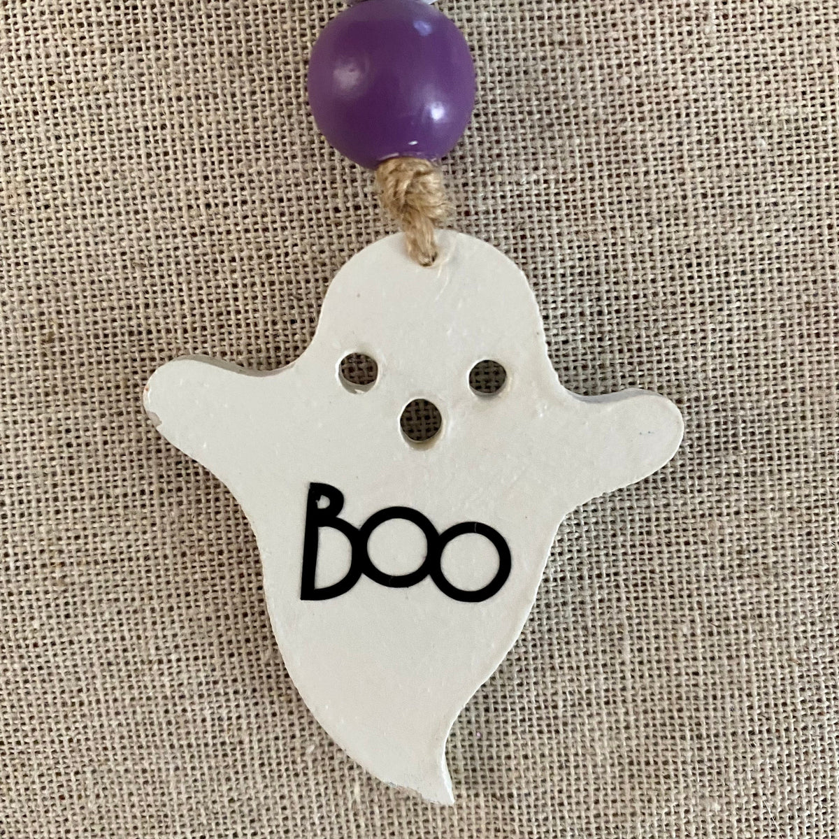 A white ghost-shaped hanging ornament with black 'BOO' lettering, two eye cutouts, and a purple bead at the top, shown against a beige burlap background.