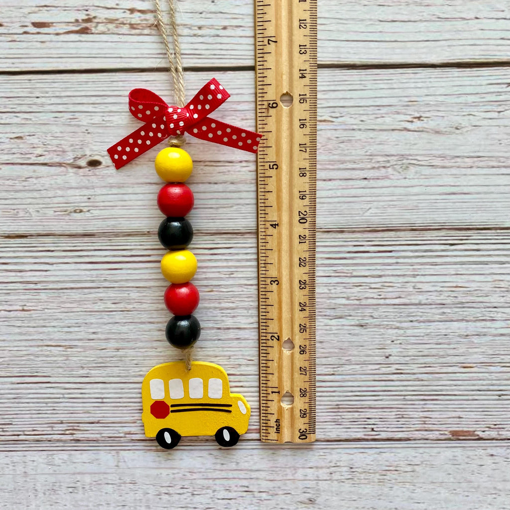 A school bus bead ornament hanging next to a wooden ruler on a whitewashed wood background. The ornament features a yellow school bus, red and black beads, a red ribbon bow with white polka dots, and a jute hanger.