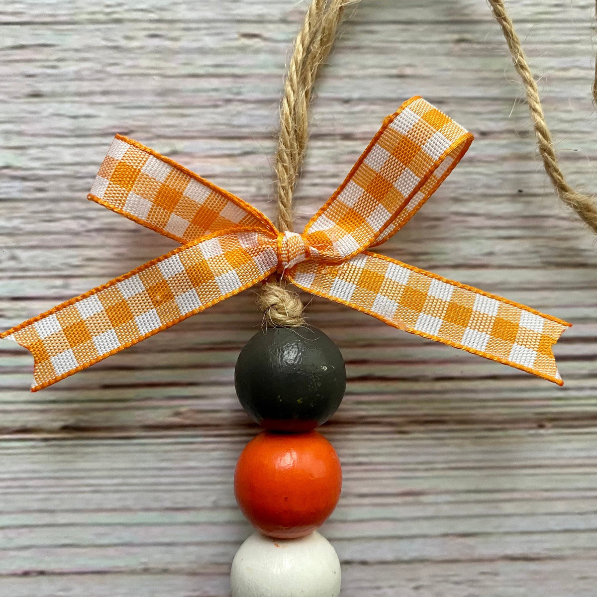 A close-up of a decorative hanging ornament with a gingham orange-and-white ribbon bow, a natural twine loop, and three stacked wooden beads in dark charcoal, orange, and white, set against a light weathered wood background.