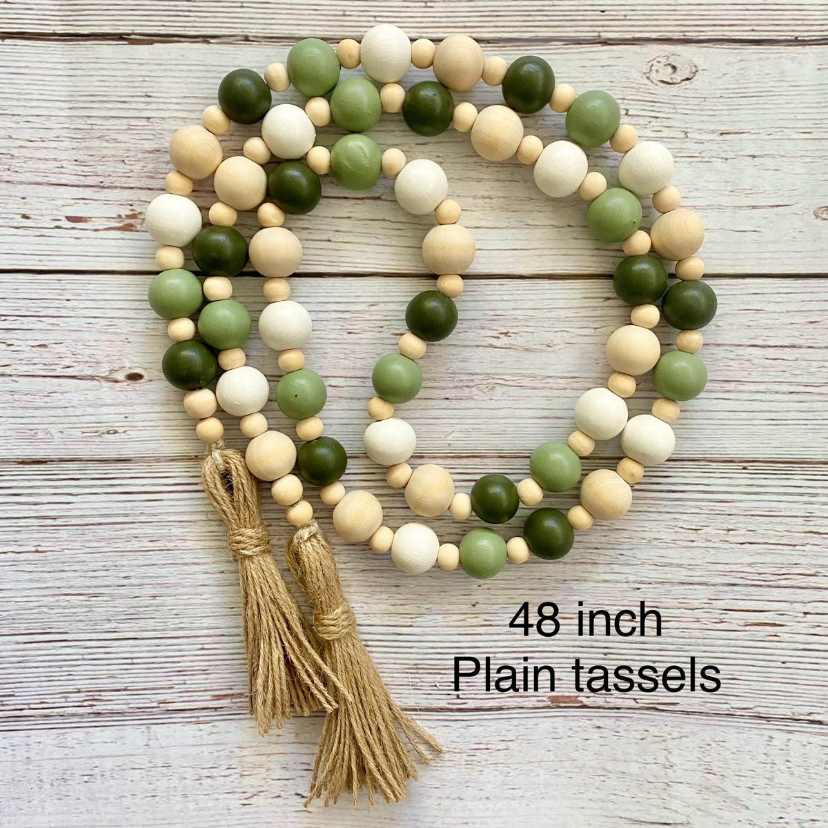 A beaded garland with alternating dark green, light sage green, cream, and natural wood beads, featuring two tan string tassels at the bottom, displayed on a whitewashed wooden background with text reading '48 inch Plain tassels'.