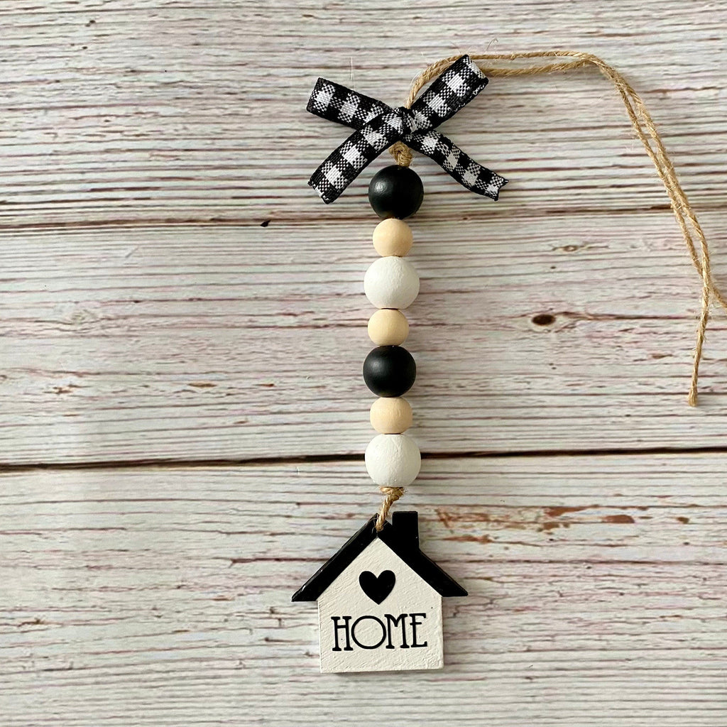 A rustic hanging ornament with a black-and-white gingham ribbon bow, natural wood beads, black beads, and a small house-shaped charm printed with the word “HOME,” displayed on a whitewashed wood plank background.