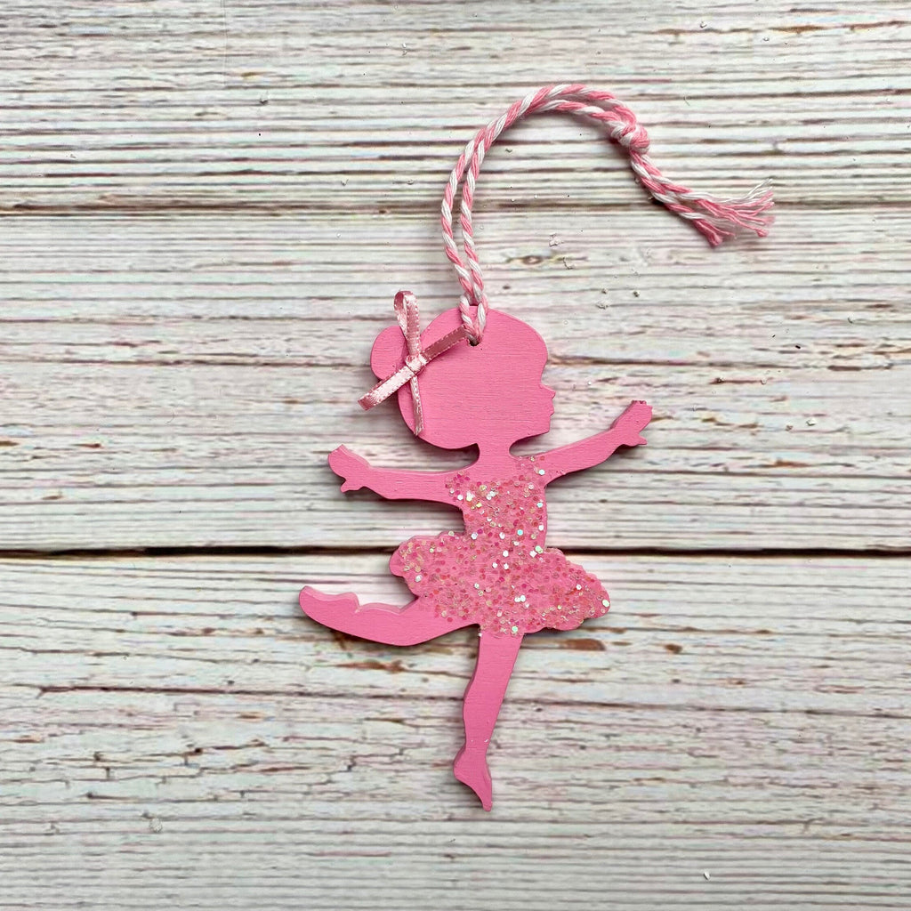 A pink ballerina silhouette ornament with glitter on the tutu and a pink-and-white twine hanging loop, displayed on a rustic whitewashed wooden surface.