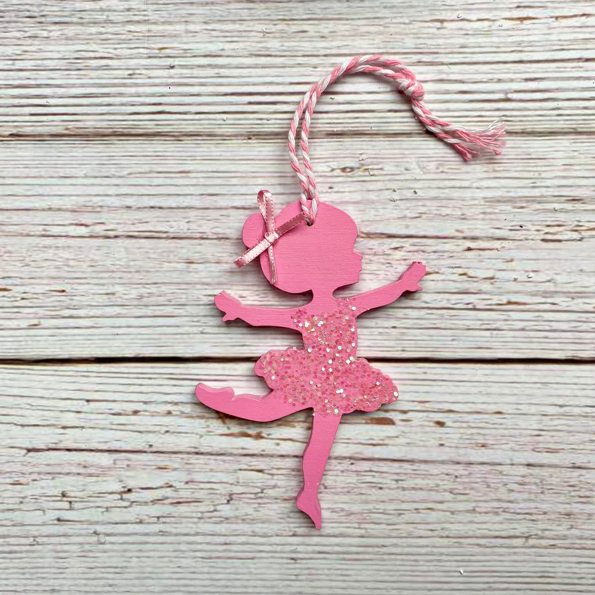 A pink ballerina silhouette ornament with glitter on the tutu and a pink-and-white twine hanging loop, displayed on a rustic whitewashed wooden surface.