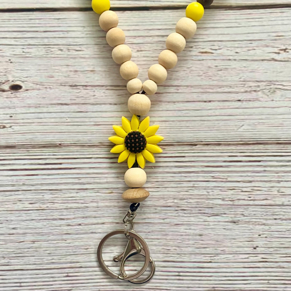 A beaded keychain or carabiner strap featuring natural wood beads, a yellow sunflower charm in the center, and a silver metal carabiner clip, displayed on a light gray wood plank background.