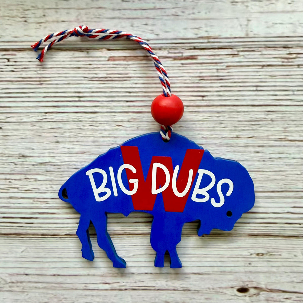 A blue wooden ornament shaped like a bison with the words "BIG DUBS" in white over a red "W" graphic, hanging from a red, white, and blue string on a whitewashed wood background.