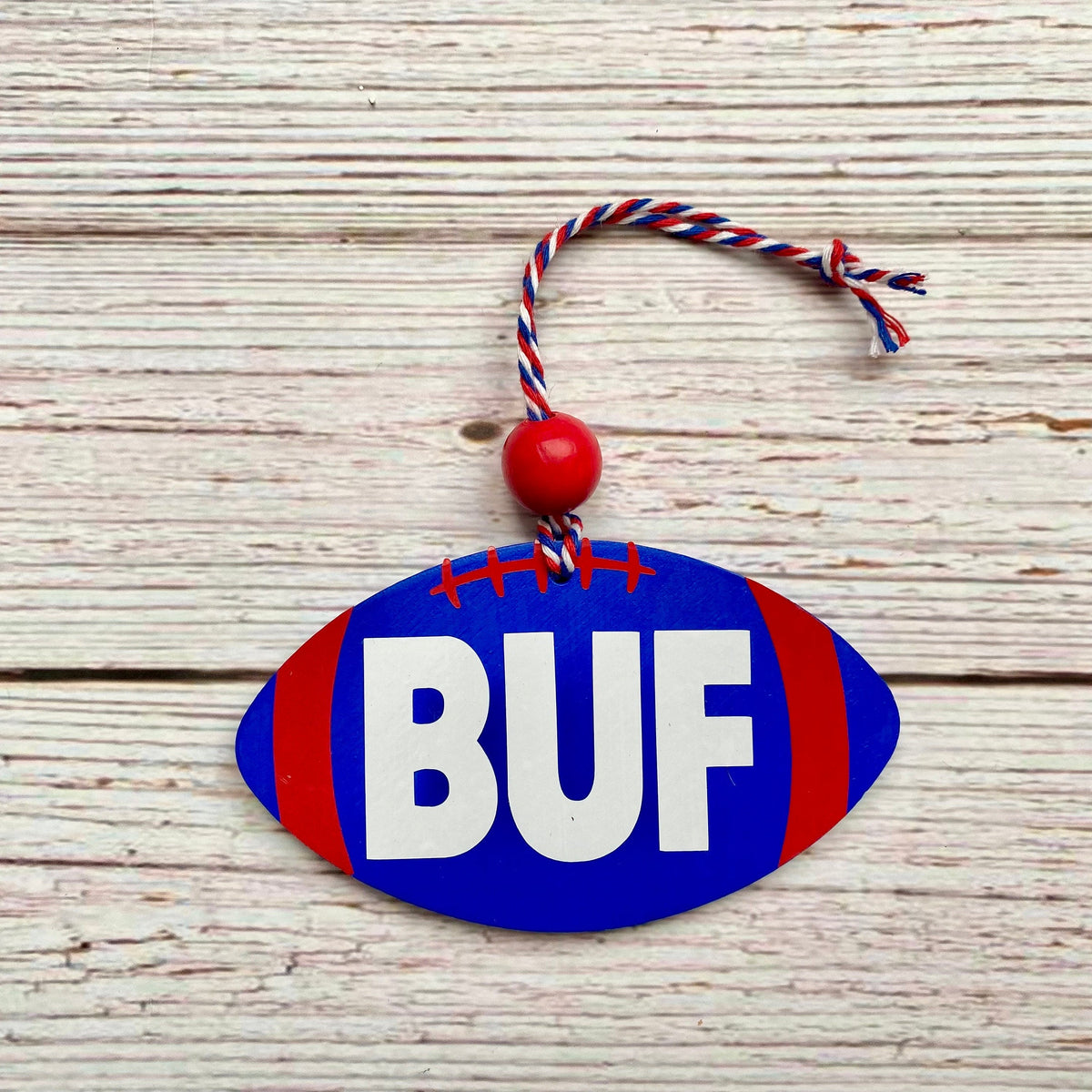 A football-shaped wooden ornament with 'BUF' in white letters on a blue background, accented with red side stripes and a red-and-white hanging string, displayed on a light wood plank surface.